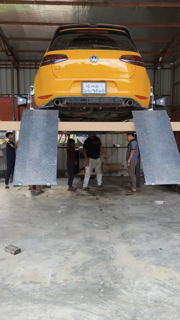 Yellow hatchback raised on a four-post lift, mechanics underneath.